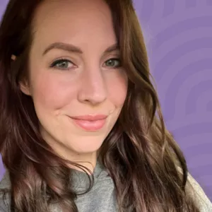 A person with long brown hair smiles slightly, wearing a grey top. The background features a lavender color with a subtle pattern.