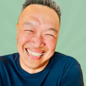 A person with short gray hair is smiling broadly, showing their teeth. They are wearing a navy blue shirt against a light green background with a subtle radial pattern.