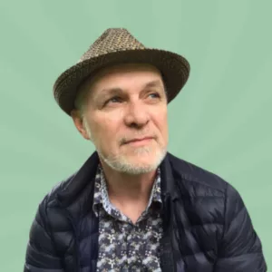 A man with a beard wearing a patterned shirt, dark jacket, and straw hat looks upward against a green, radial gradient background.