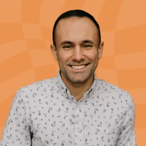 A person with short dark hair, smiling and wearing a light-colored button-up shirt with small patterns, stands in front of an orange wavy background.