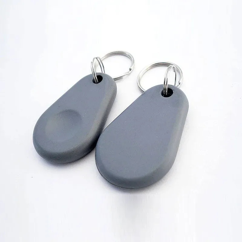 Divi galvenie FOB NFC pelēkā krāsā, ar metāliskiem atslēgu gredzeniem un NFC tehnoloģiju, Atpūtieties uz balta fona.