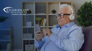 An older man wearing a blue button-down shirt, black eyeglasses, and white headphones sits in his living room, leaning back in his chair and scrolling on his phone. The image has a blue shadow overlay with the Florida Eye Specialists logo in the top left corner