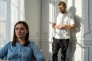 Forgiveness in Relationships: How to Let Go of Anger & Resentment 4 A stock photo showing a white man and woman in a sunny room with white walls. The woman is sitting in the foreground and the man is standing against the wall behind her. They both look serious.