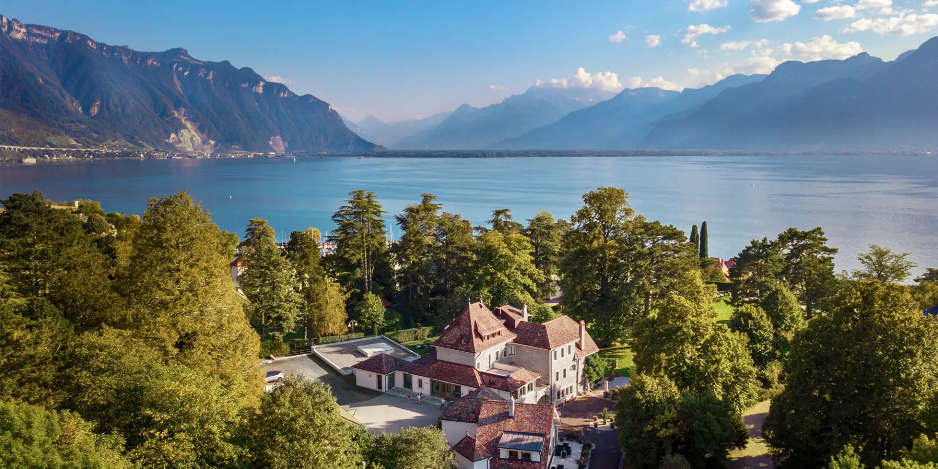 LEE_About_1 Aerial view of a GuestLee luxury rental in Switzerland, nestled by trees, lake, and mountains under a blue sky. | GuestLee luxury rental Switzerland