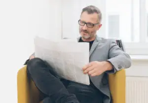 LEE_NewPaper Man with glasses relaxes in a bright, stylish Geneva GuestLee rental, reading a newspaper in a luxe armchair by the window. | GuestLee luxury rental Switzerland