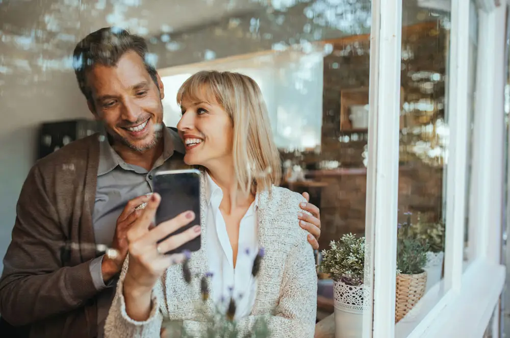 LEE_Owner Smiling couple in a luxury GuestLee Geneva villa browse rentals on smartphone near bright window with potted plants. | GuestLee luxury rental Switzerland