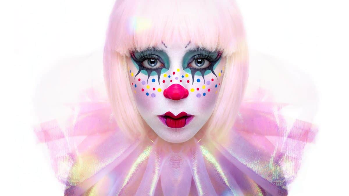 Person with white-pink hair, bright clown makeup with colorful dots, exaggerated eyes, and a ruffled pink collar, staring directly at the camera against a white background.