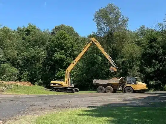 An excavator with an extended arm is loading dirt into a dump truck on a paved area near a wooded landscape, demonstrating expert site preparation and grading services. Kenneth Bros