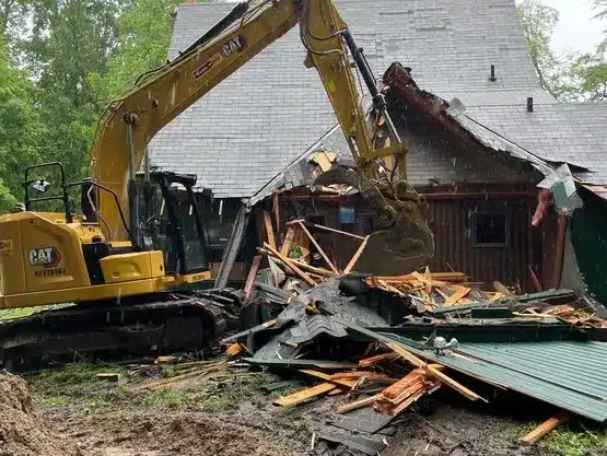 A yellow excavator demolishes part of a wooden house, with debris scattered on the ground—expert work from a leading commercial demolition contractor in mid Michigan. Kenneth Bros