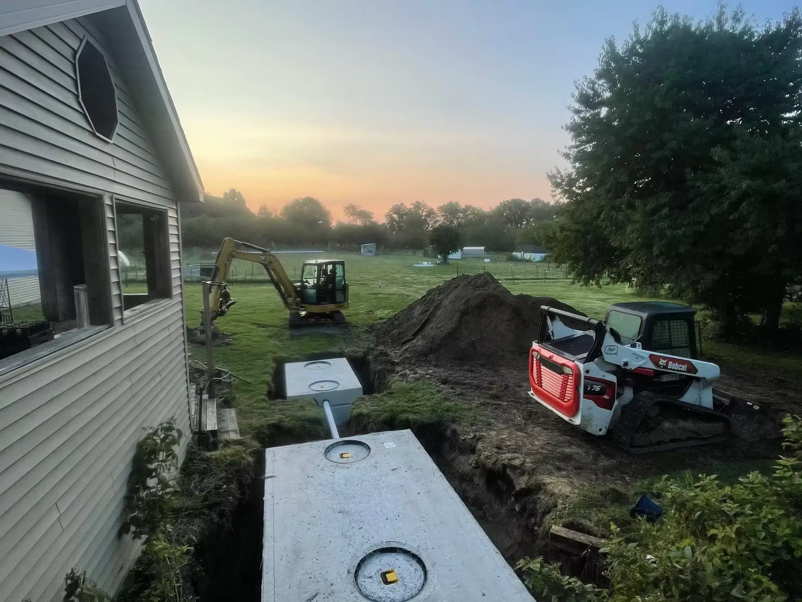 Excavation of a yard next to a house with construction equipment and newly installed concrete septic tanks visible in the ground, showcasing expert septic system installation Michigan and site preparation and grading services. Kenneth Bros