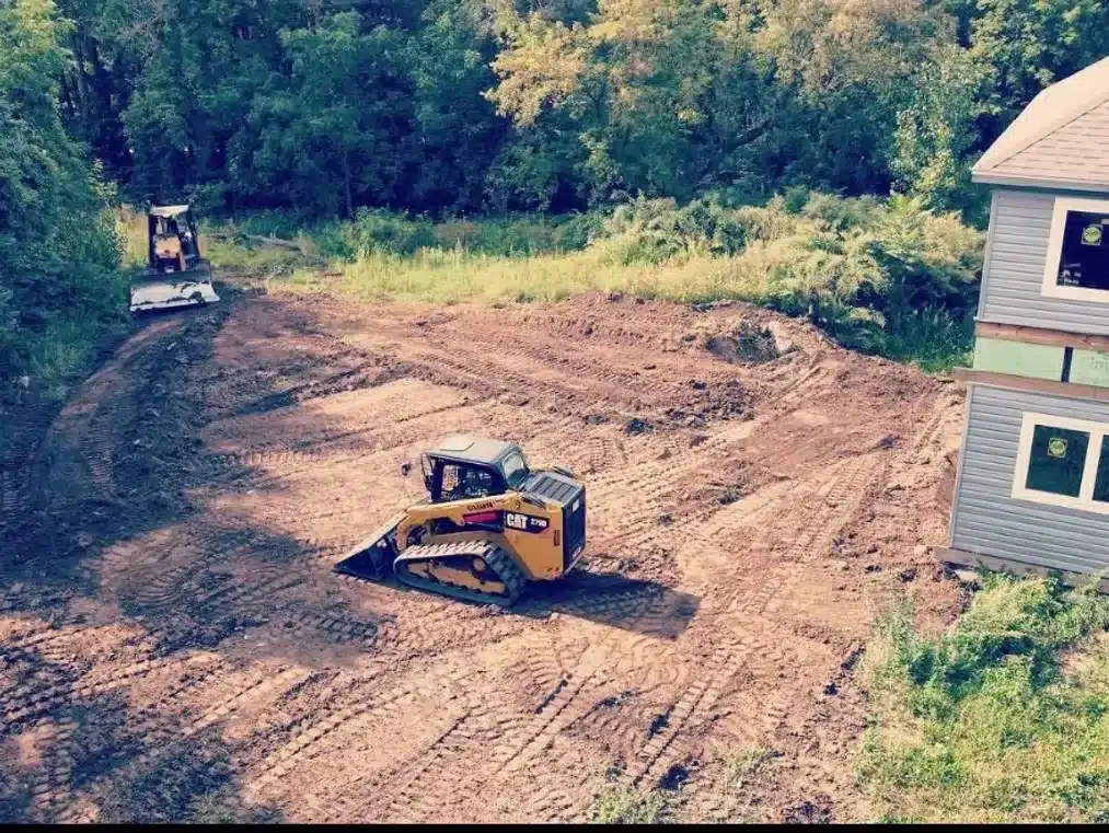 A yellow bulldozer levels dirt near a house under construction, while excavation contractors DeWitt MI work with another vehicle in the background surrounded by trees. Kenneth Bros