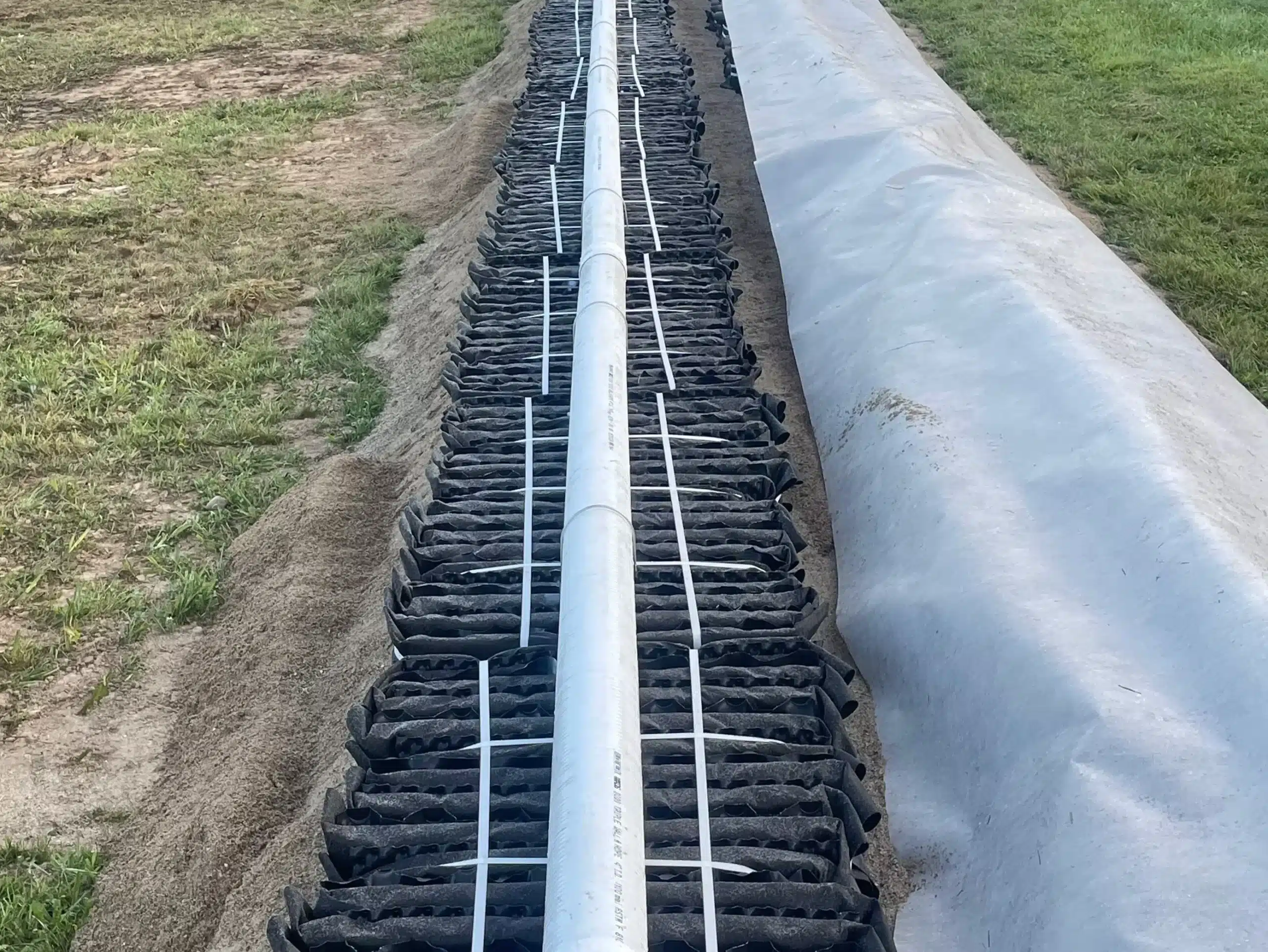 A long PVC pipe is laid over rows of black geogrid material and soil, part of septic system installation Michigan, with a sheet of plastic covering the ground to the right. Kenneth Bros