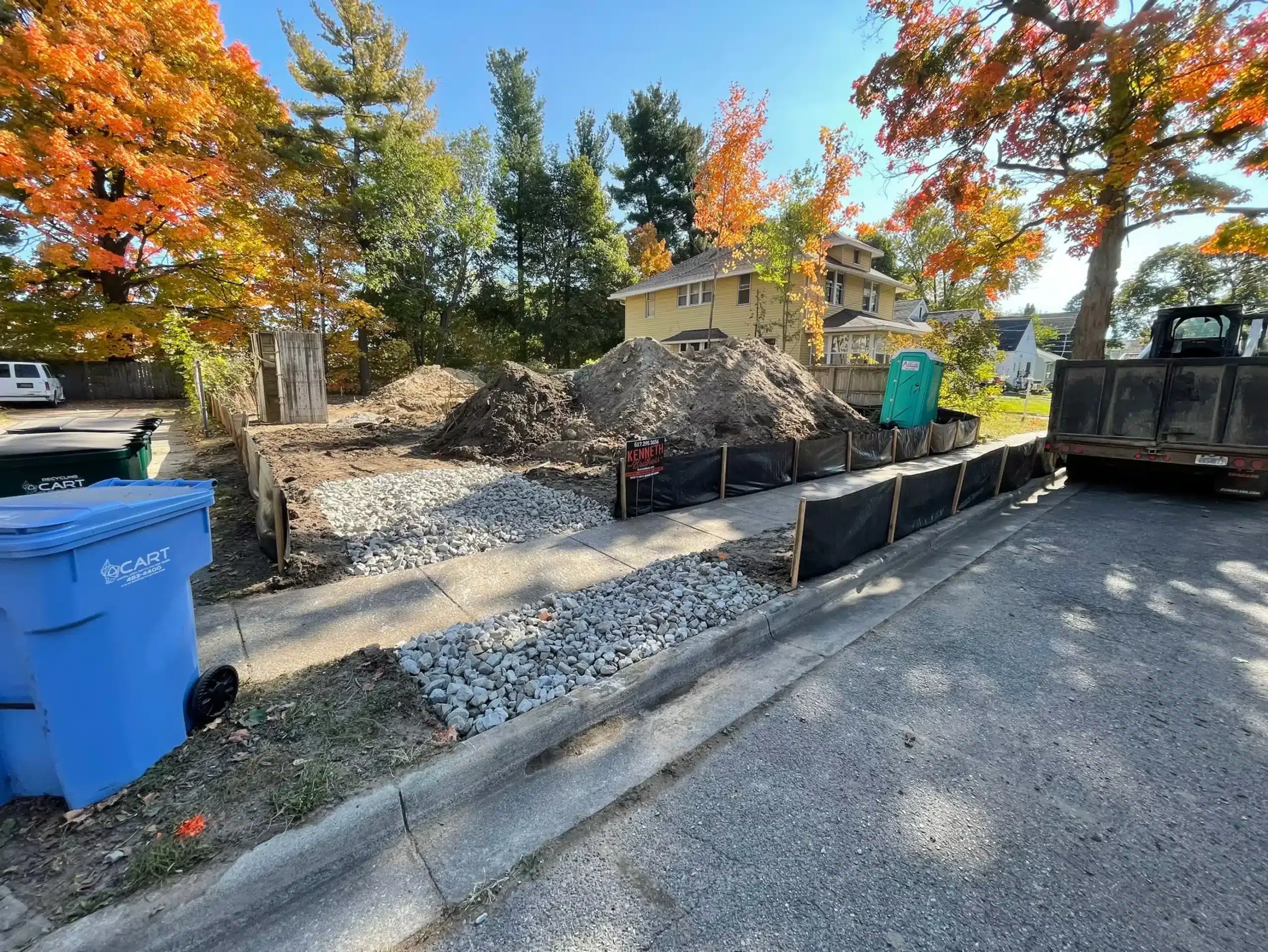 A residential construction site with gravel and dirt piles, black barriers, a portable toilet, and a blue recycling bin on a tree-lined street showcases site preparation and grading services in action. Kenneth Bros