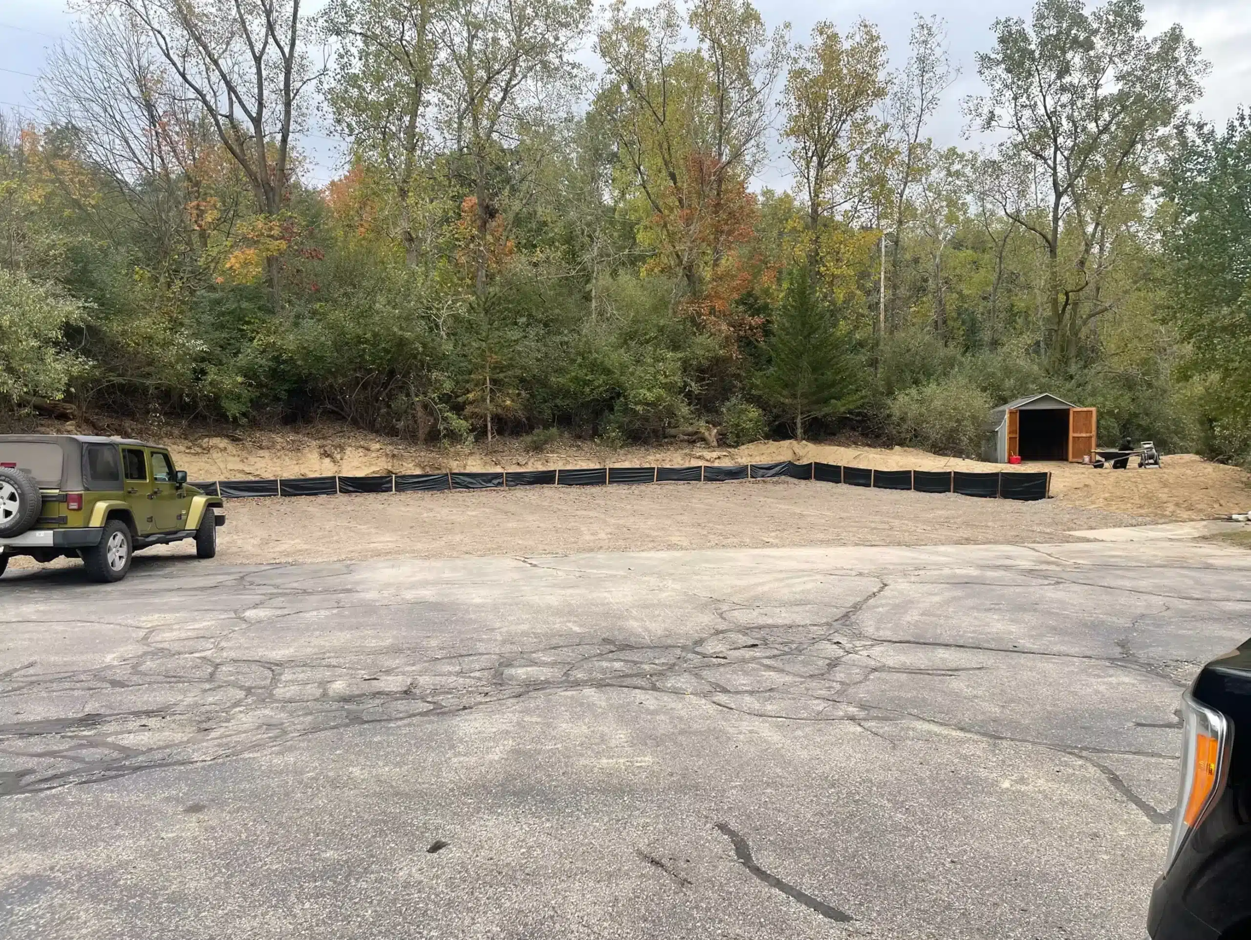 A fenced-off rectangular area filled with dirt is beside a wooded area; perfect for site preparation and grading services. A Jeep, a shed, and part of another vehicle are visible in the foreground and background. Kenneth Bros