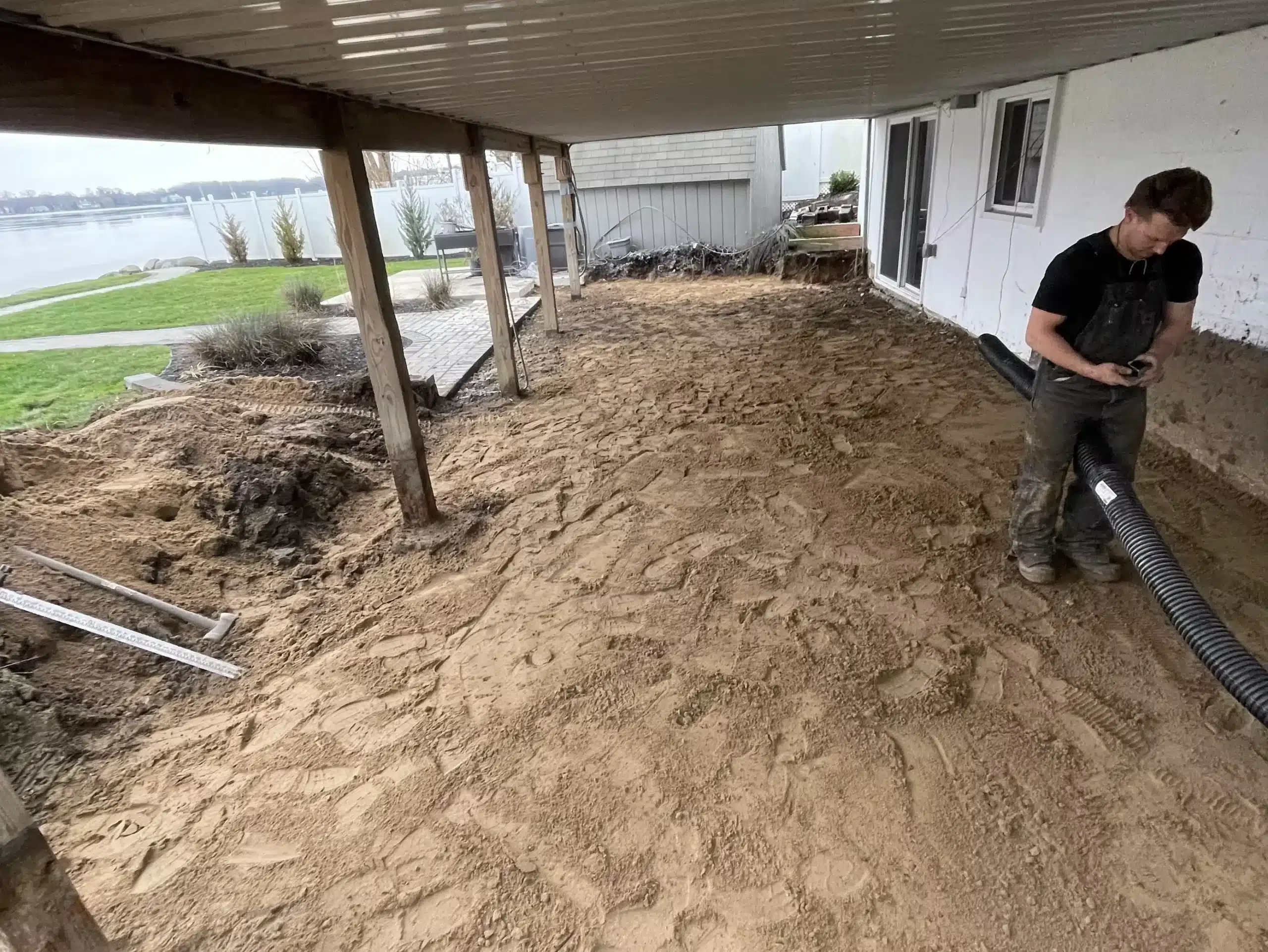 A person stands on a sandy surface under a covered patio next to a house, holding a flexible hose. The area, possibly being prepped by excavation contractors DeWitt MI, appears to be under renovation or landscaping. Kenneth Bros