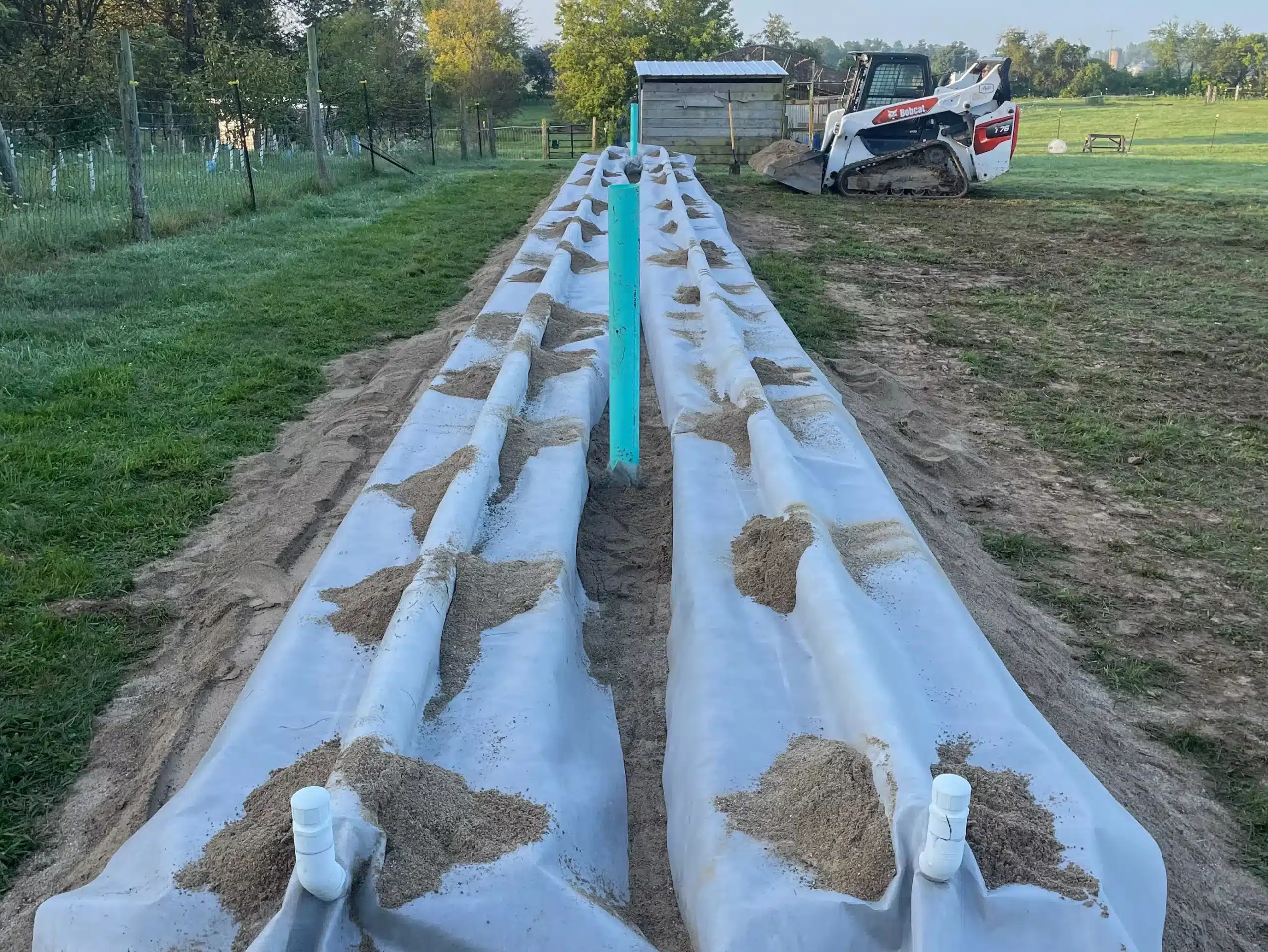 Two long white plastic grow beds with soil mounds are set up outdoors on grass, with a small building and construction vehicle in the background—ideal for projects managed by a commercial demolition contractor mid Michigan. Kenneth Bros