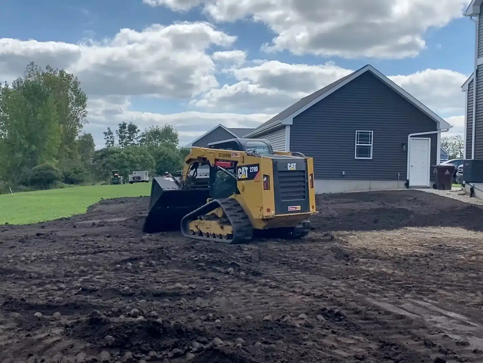 A yellow Caterpillar skid steer loader, operated by leading excavation contractors DeWitt MI, levels dirt in the yard of a house with dark gray siding under a partly cloudy sky. Kenneth Bros
