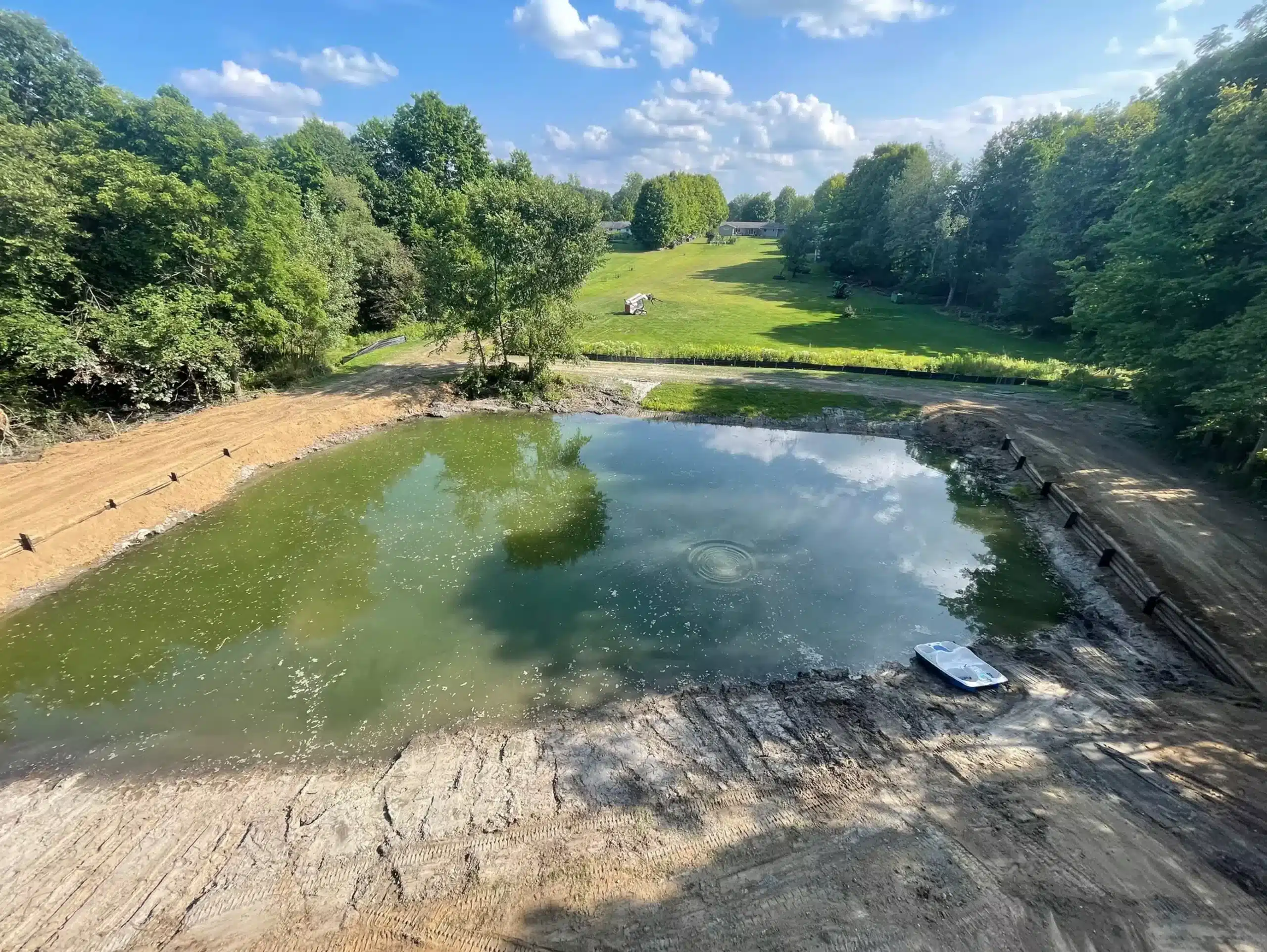 A small, partially drained pond with a rowboat on the shore—possibly a site visited by excavation contractors DeWitt MI—surrounded by trees and grassy fields under a mostly clear sky. Kenneth Bros