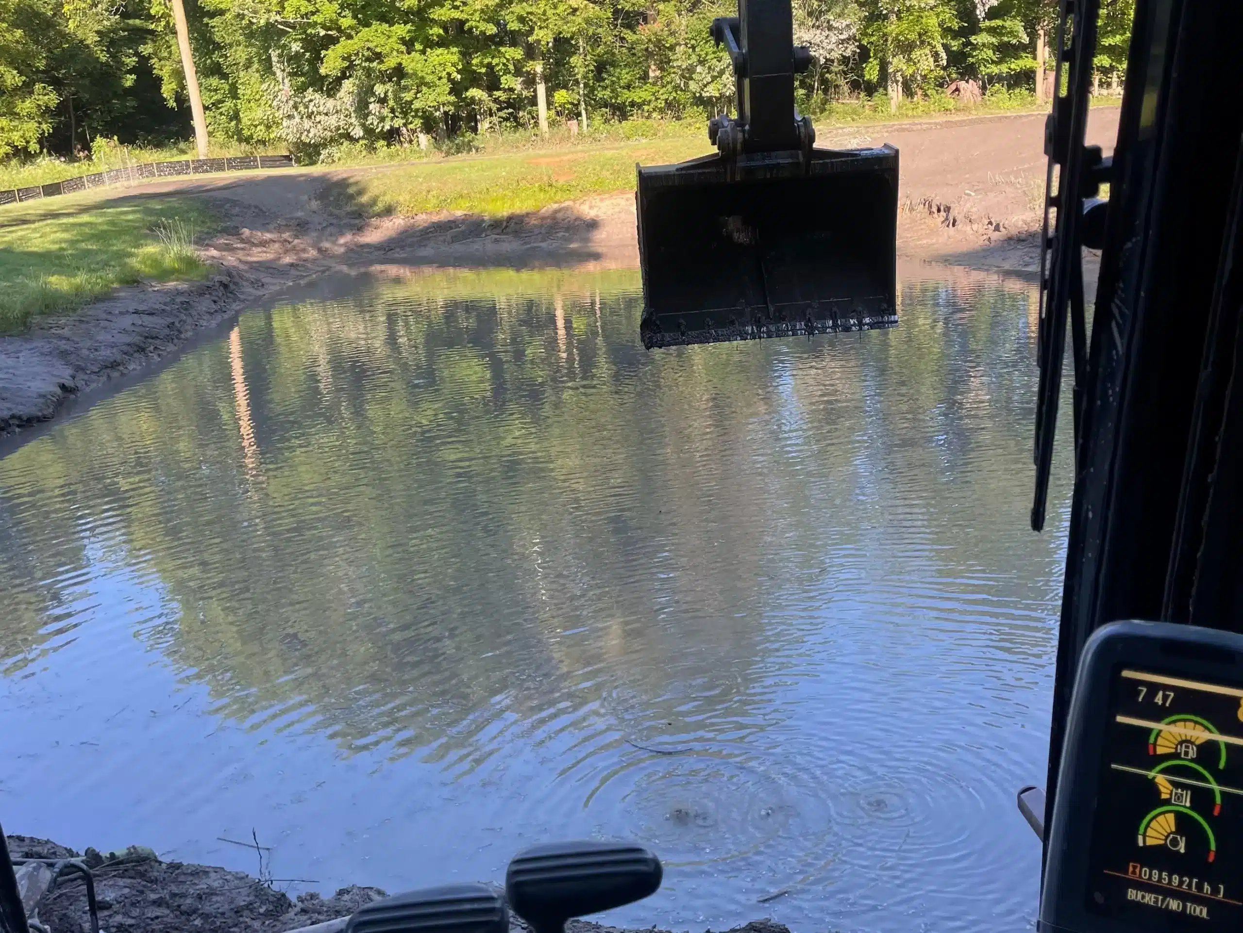 A view from inside an excavator cab overlooking a pond, with the excavator bucket lifted above the water and trees in the background—showcasing excavation contractors DeWitt MI at work in a picturesque setting. Kenneth Bros