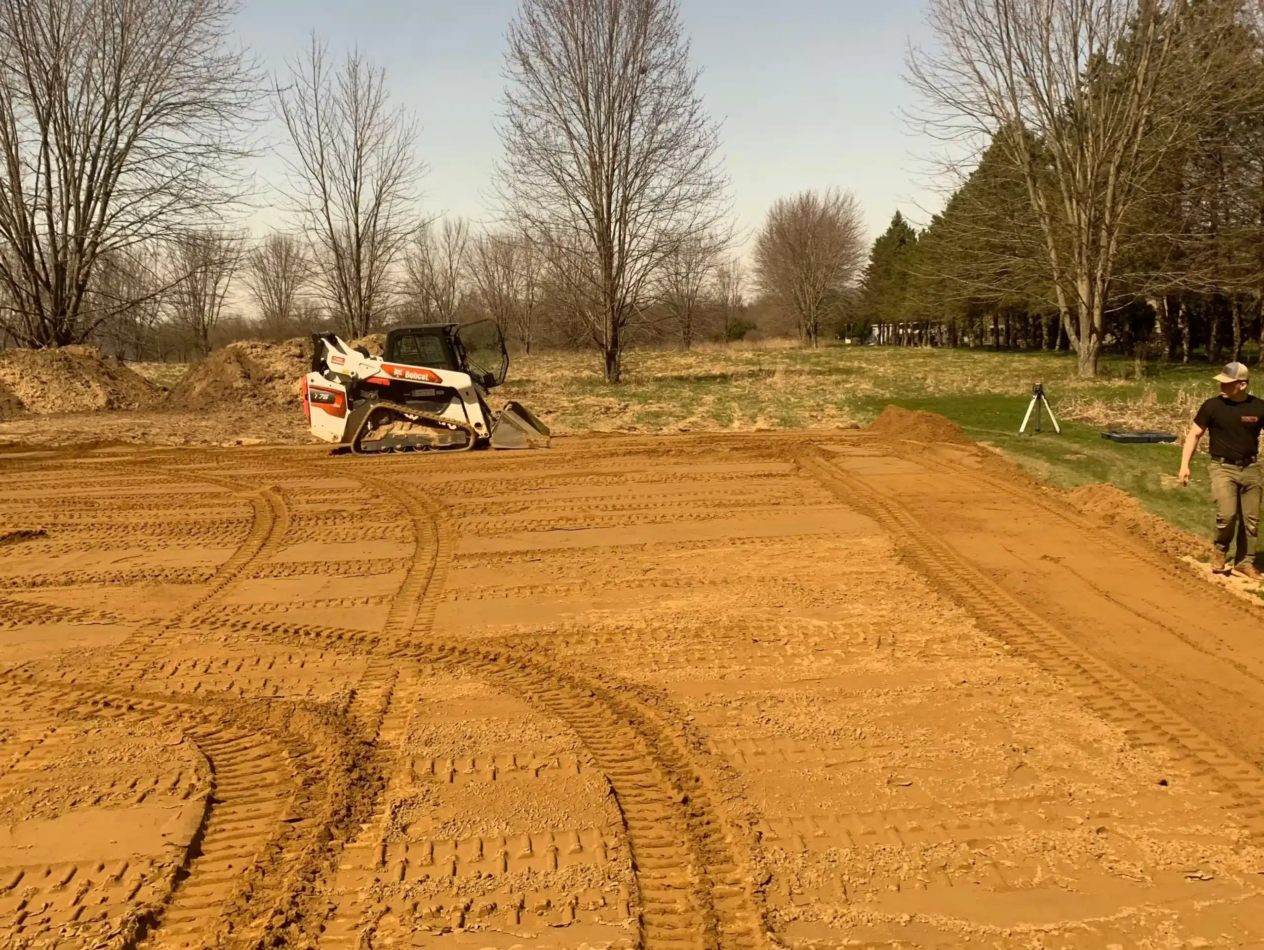 A skid steer loader and a person stand on a leveled dirt area with visible tire tracks; trees and grass surround the site under a clear sky—a scene typical for excavation contractors DeWitt MI. Kenneth Bros