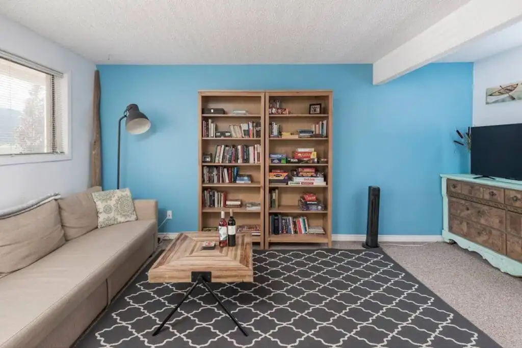 A living room perfect for a vacation rental, featuring a beige sofa, wooden coffee table, two bookshelves on a blue wall stocked with board games, and a TV on a turquoise dresser. A patterned rug covers part of the floor.