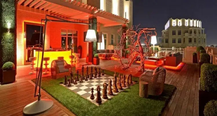 Rooftop terrace at night with a large outdoor chessboard, wicker chairs, modern lighting, a bar area, illuminated decorative plants, and vacation rental board games for extra fun.