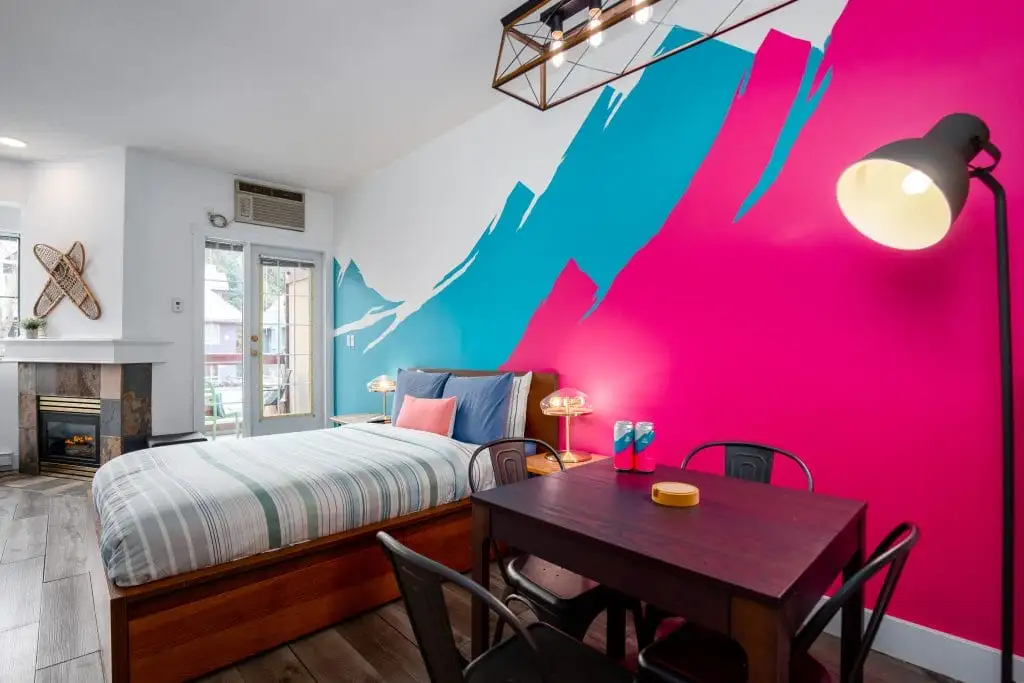 A modern bedroom designed for buying a vacation rental, with a striped bed, dining table, black chairs, wall lamp, and a bold pink and blue mural. A fireplace and glass door complete the inviting atmosphere.