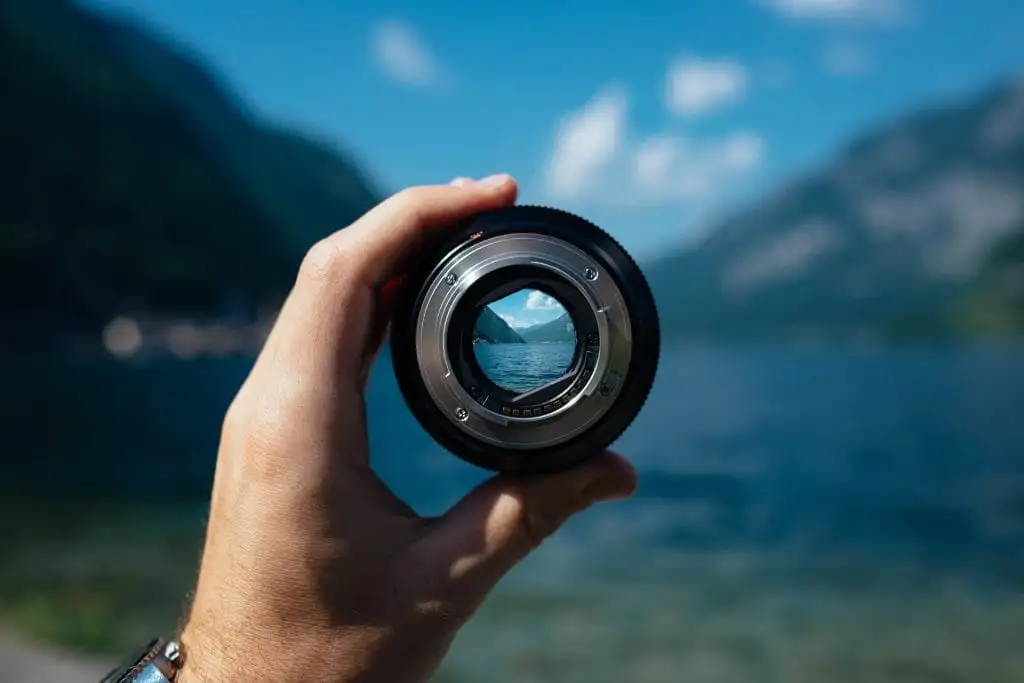 A hand holds a camera lens with a scenic view of mountains, lake, and sky—perfect inspiration for things to do in Sunshine Valley—visible through the glass, while the background remains softly blurred.
