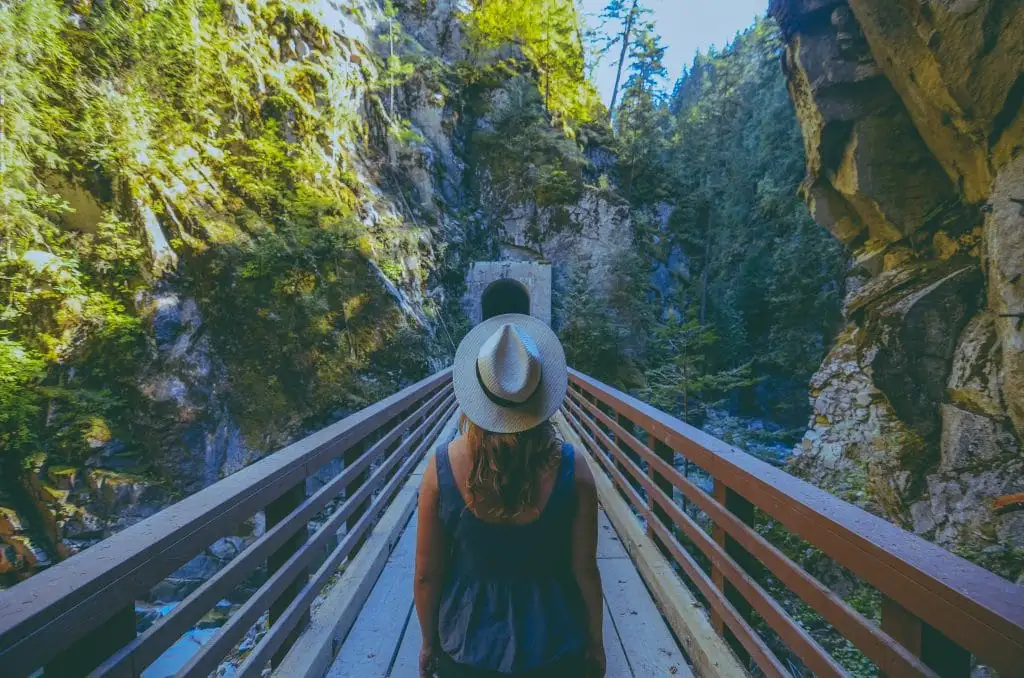 A person wearing a hat and backpack stands on a wooden bridge in a forested canyon, facing away toward a tunnel in the distance—one of the scenic things to do in Sunshine Valley.