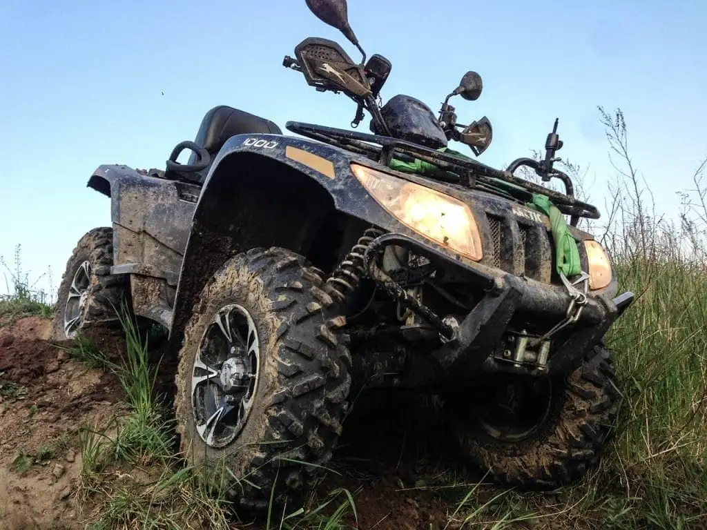 A muddy ATV with headlights on is parked on a grassy slope, facing slightly upward under a clear sky—capturing the spirit of adventure among the top things to do in Sunshine Valley.