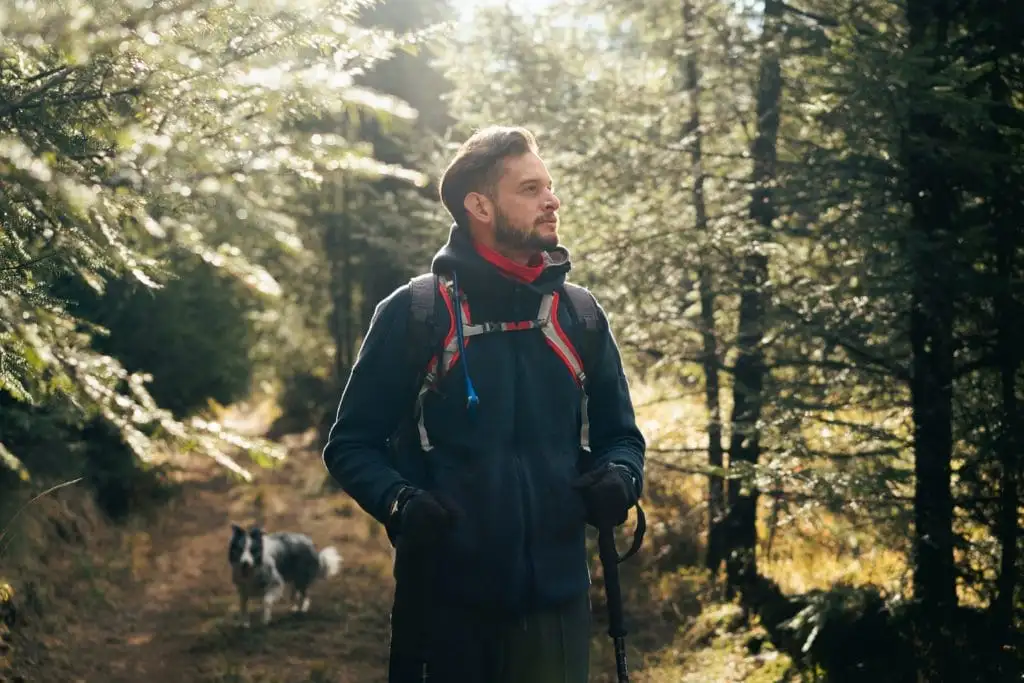 A man wearing hiking gear walks through a sunlit forest trail with a dog following behind him, enjoying the peace often found near top vacation rental management retreats.