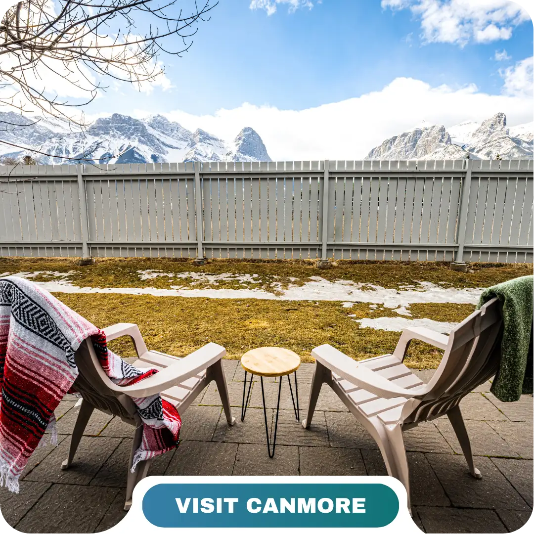 Two Adirondack chairs with blankets face a small table on a patio, overlooking a fenced yard with snow patches and snow-capped mountains in the background. Experience Canmore with Lifty Life.