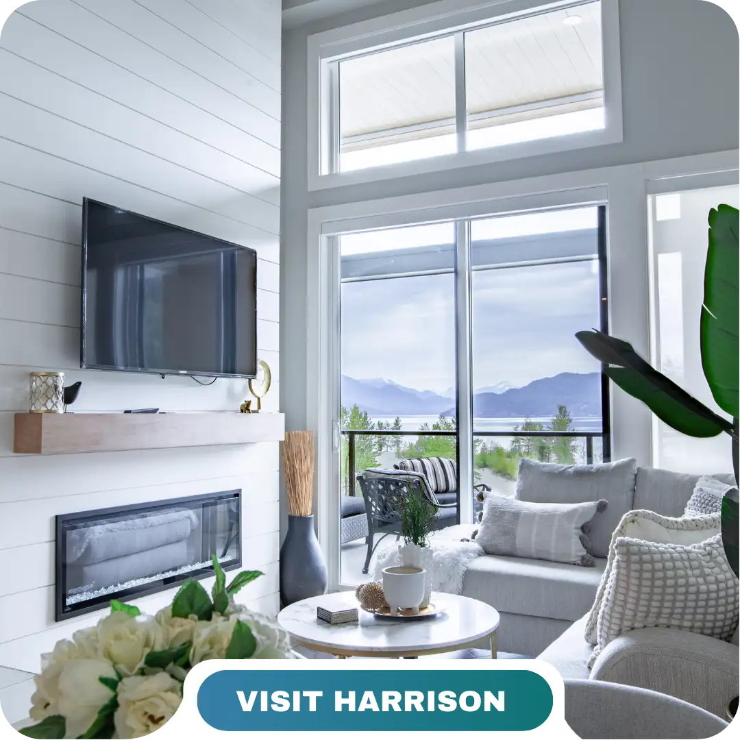Modern living room with a fireplace, wall-mounted TV, and large windows showcasing scenic mountain and lake views. Experience the Lifty Life—tap the "Visit Harrison" button below to explore further.