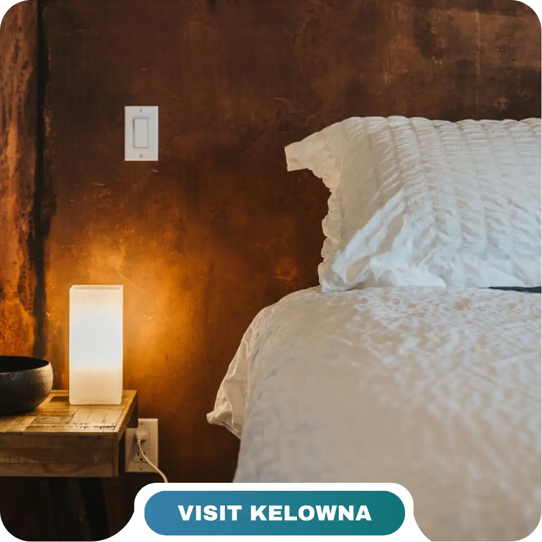 A bedside table with a lit rectangular lamp, a bowl, and a bed with white bedding against a brown textured wall. A switch is seen above the table. Text reads "Visit Kelowna with Lifty Life.