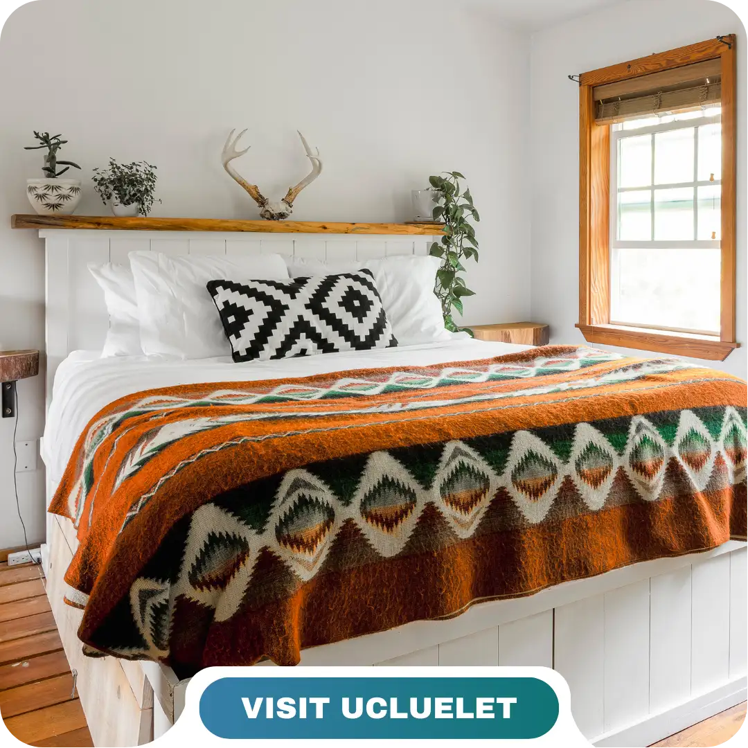 A neatly made bed with white linens, an orange patterned blanket, and a black-and-white cushion in a bright, rustic room; text reads "VISIT UCLUELET with Lifty Life.