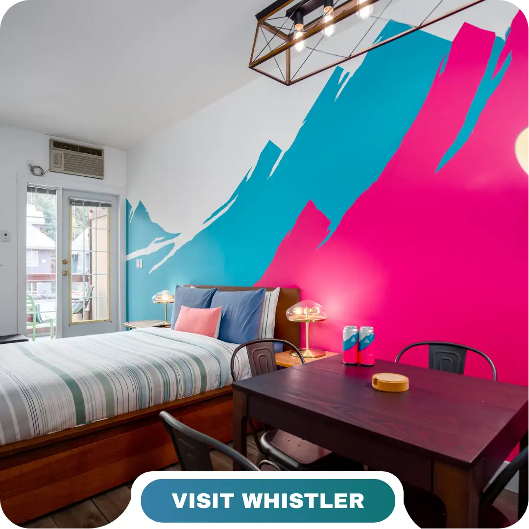 A modern bedroom with a striped bed, wall art of blue and pink mountains, a small dining table, and a "Visit Whistler" label at the bottom—perfect for enjoying Lifty Life comfort.