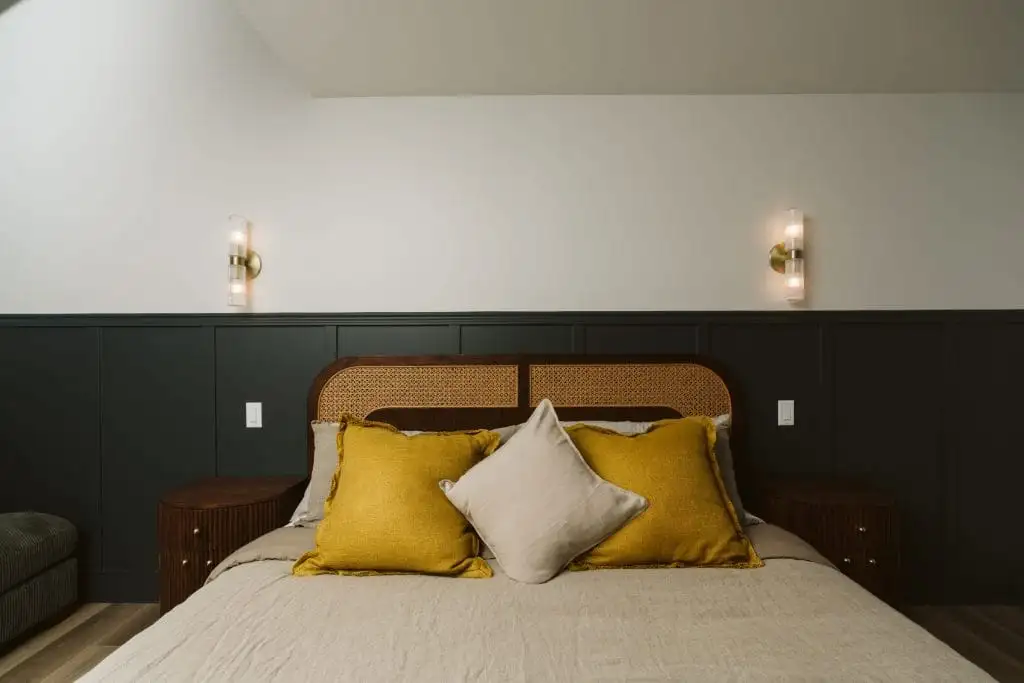 A neatly made bed with mustard yellow and beige pillows against a dark wainscoted wall, flanked by two wall-mounted lights and dark wooden nightstands, offers a cozy glimpse into our “about us” style of thoughtful interior design.