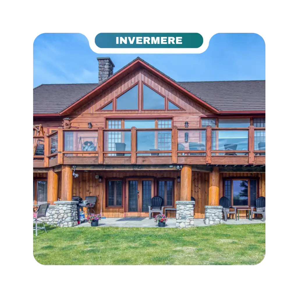 A two-story wooden house with large windows, stone pillars, a balcony with glass railing, patio chairs, and potted flowers on a grassy lawn is perfect for homeowners seeking beautiful destinations to unwind.