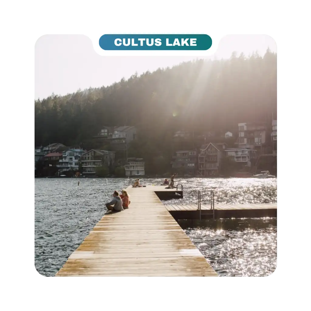 A wooden dock extends into Cultus Lake with guests sitting along the edge; houses and trees line the background under sunlight, making it one of the area's inviting destinations.