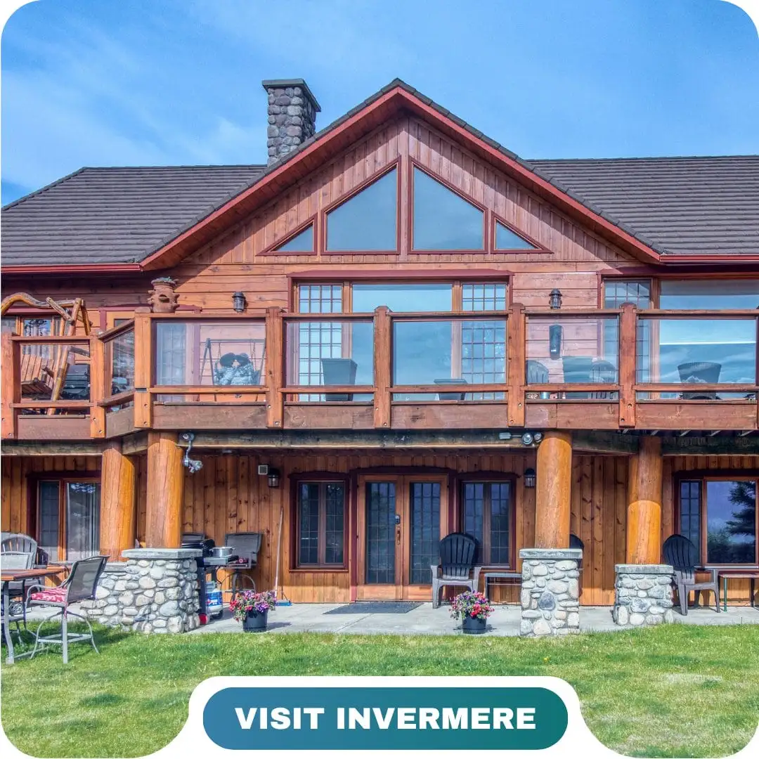 A wooden lodge-style Lifty Life house with large windows, a balcony, and stone pillars, set on a grassy lawn under a clear blue sky. Text reads "VISIT INVERMERE.