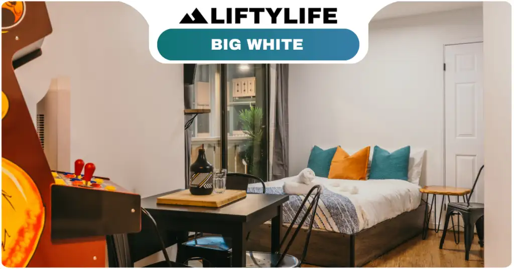 A cozy studio room with a bed, colorful pillows, small dining table, chairs, and an arcade machine; perfect for those interested in buying property or exploring real estate at Big White. A window and door sit beneath a “Liftylife Big White” banner.