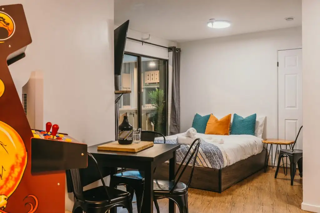 A cozy studio with a bed, a small dining table for four, an arcade machine, and a wall-mounted TV; wooden floors and a window overlooking greenery—perfect for guests seeking vacation rental management in Big White.