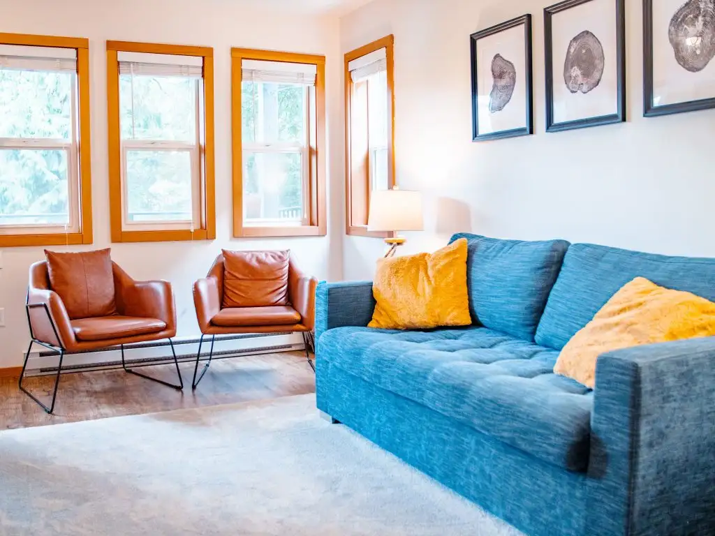 A bright living room with three windows, two brown chairs, a blue sofa with yellow pillows, a lamp, and framed art on the wall—perfect for vacation rental management in Ucluelet.