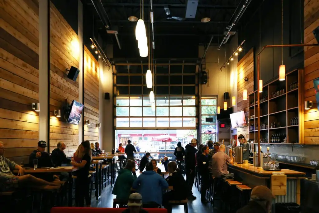 People sit at tables and the bar inside a modern, high-ceilinged brewery with wood-paneled walls, pendant lights, and large windows—one of the top Things to Do in Big White for those looking to relax with friends and craft beer.