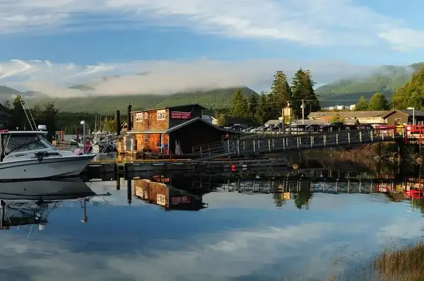 A wooden building and dock are reflected in calm water, with boats moored nearby—a serene scene that captures the natural beauty one can experience when buying property in Ucluelet, set against a forested, mountainous backdrop under a partly cloudy sky.