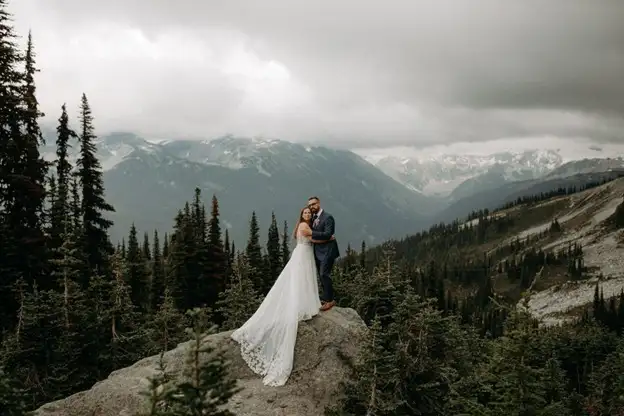A bride and groom stand on a large rock surrounded by pine trees, mountains, and clouds in the background—capturing the magic of a Wedding in Whistler.