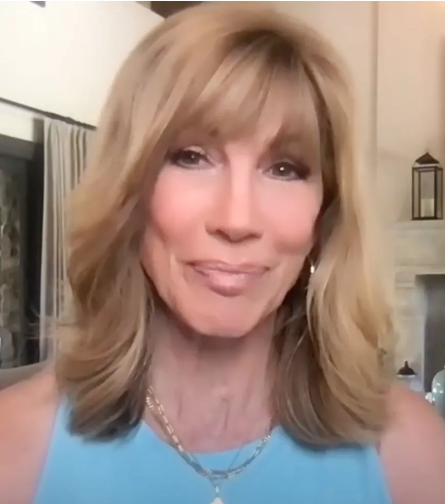 A woman with shoulder-length blonde hair and bangs smiles softly at the camera, wearing a light blue top and layered gold necklaces. The softly lit, modern room behind her reflects her passion for giving and helping others through charity.