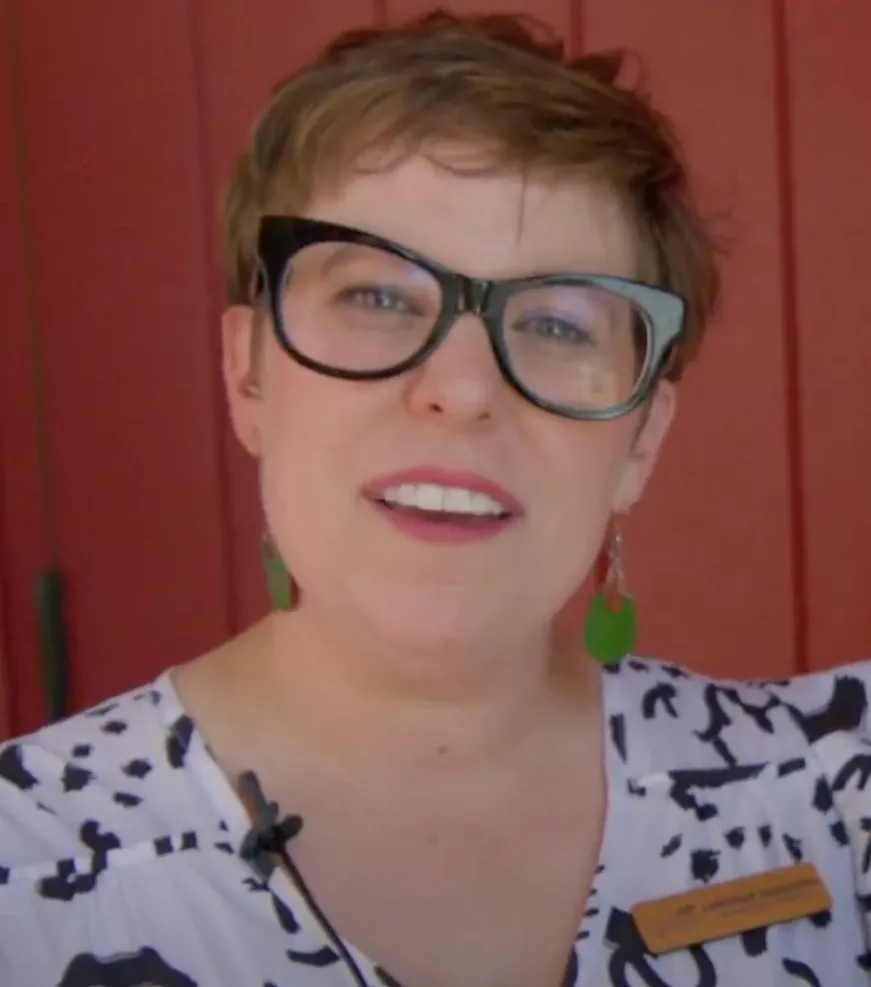 A woman with short brown hair, large black glasses, and green earrings smiles at the camera. Wearing a white and black patterned top and a name tag, she stands in front of a red wall, embodying the spirit of giving and charity.