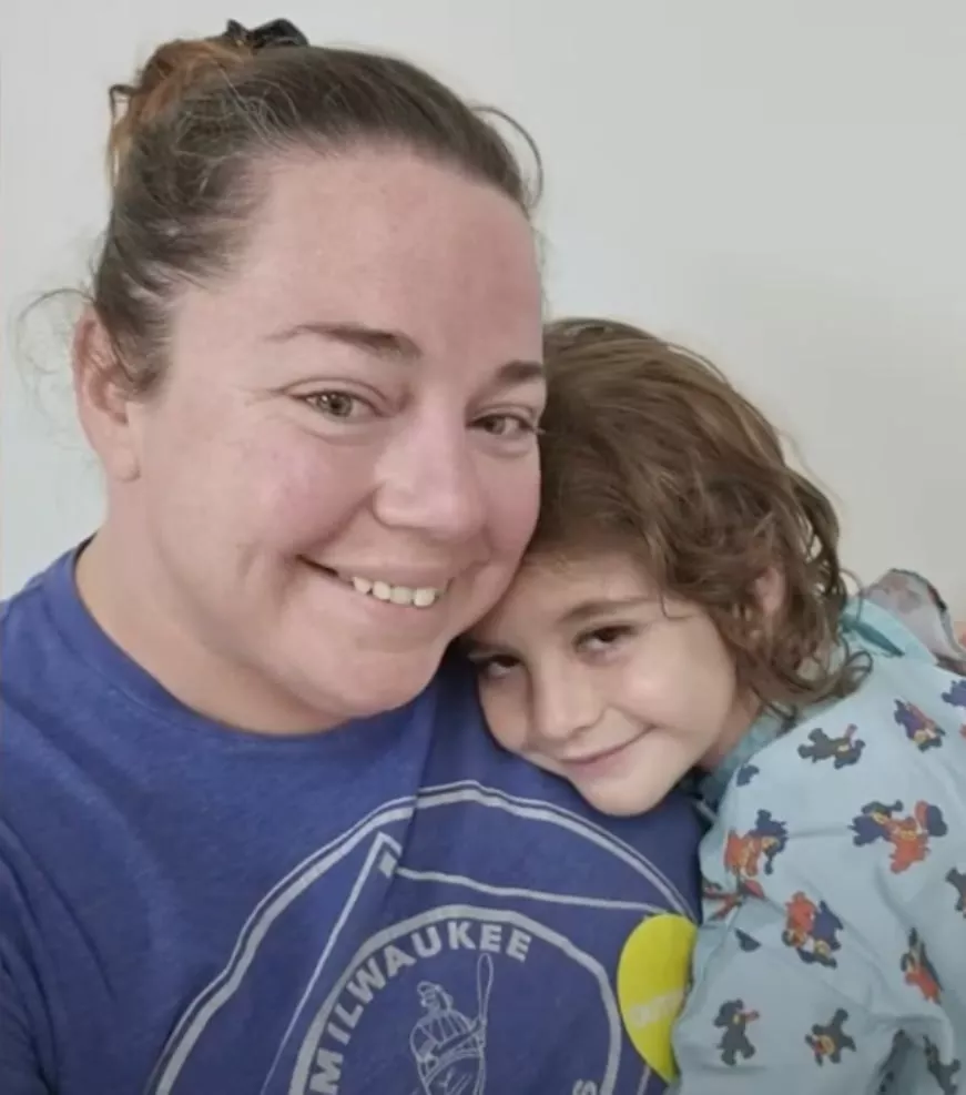 A woman with brown hair in a ponytail smiles as a young child in printed pajamas hugs her, both looking at the camera. Their warm embrace captures a moment of giving and help against a plain light background.