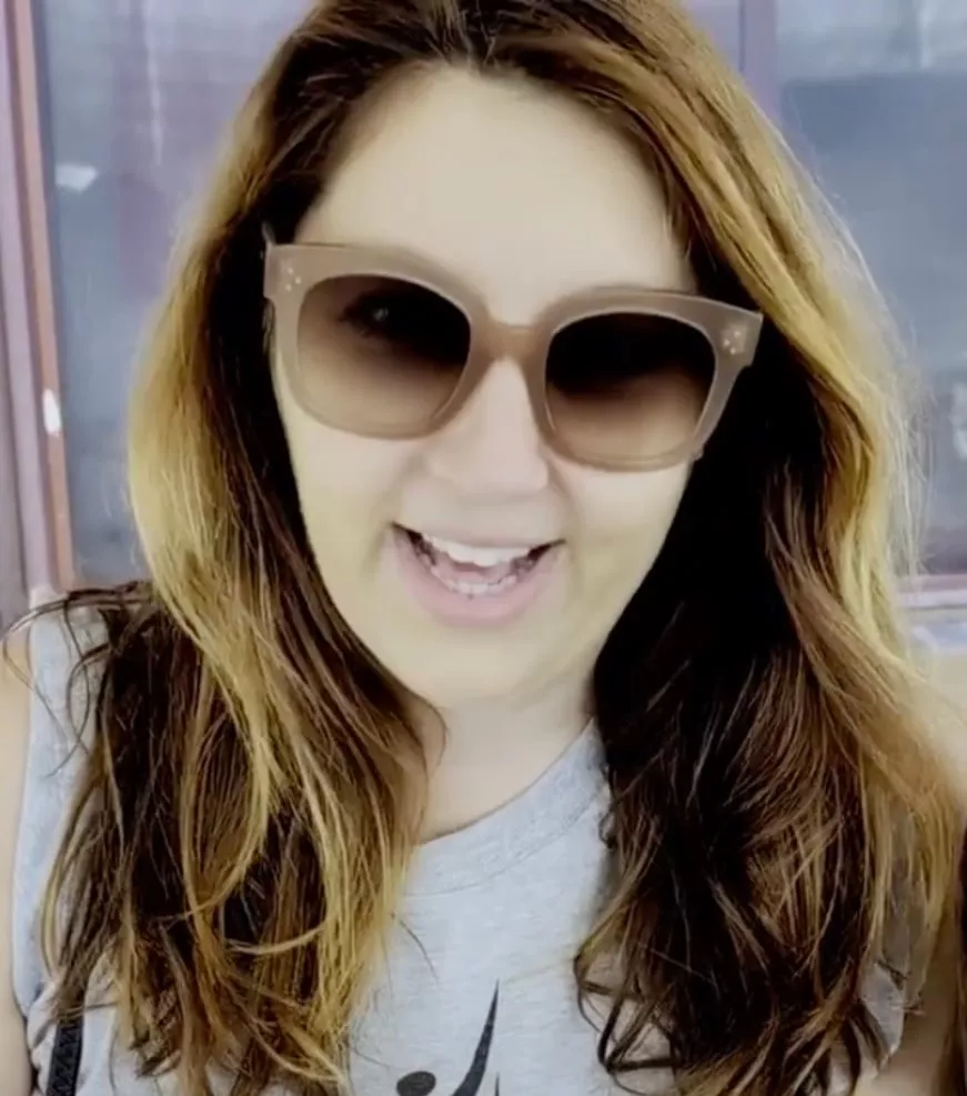A woman with long brown hair wearing large sunglasses and a sleeveless gray top smiles while standing outside in front of a window, radiating the warmth of giving.